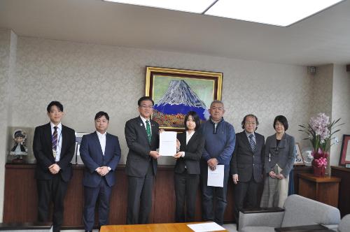 富士山の絵のある応接室で、中央に市長と一緒に紙を持って立つ女性と、その周囲にスーツの男女5名