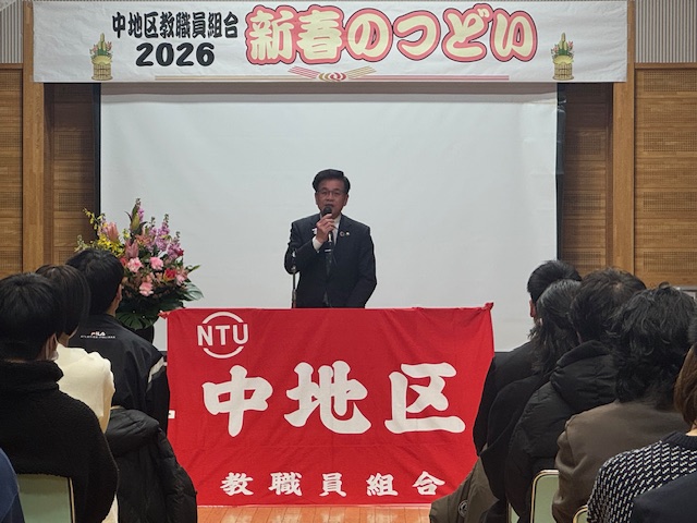 中地区教職員組合の赤い旗が貼られた演台の後ろに立ってマイクを持って挨拶する市長