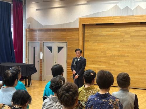 広い部屋でスタンドマイクの前で挨拶する市長と、それを聞く着物姿の女性9人