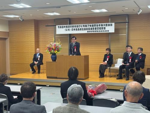 低いステージで演台の後ろに立って挨拶する市長とその右側にスーツの来賓4名が座る。