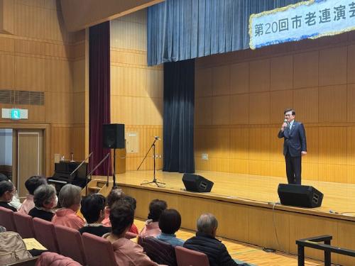 ステージの上で立ってマイクを持ち挨拶する市長と、手前に座っている観客12人ほど。