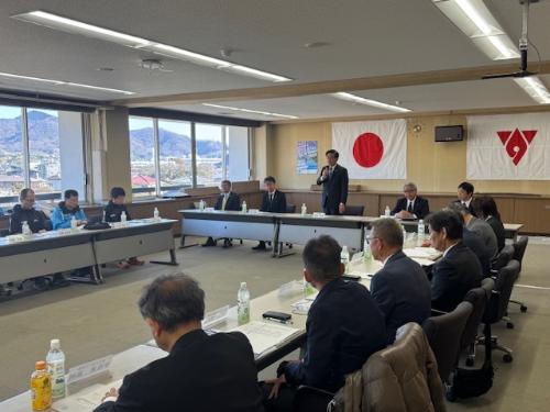 会議室でスーツの男性が座っている。奥に市長が立って挨拶をしている。その後ろの壁には国旗と市旗が掲げられている。