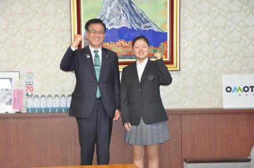 富士山の絵のある応接室で、市長とガッツポーズをする高校の制服を着た女性。