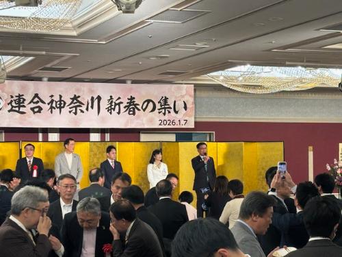 連合神奈川新春の集いの看板と金屏風があるステージで挨拶する市長と、左側に来賓4名、手前に出席者多数。