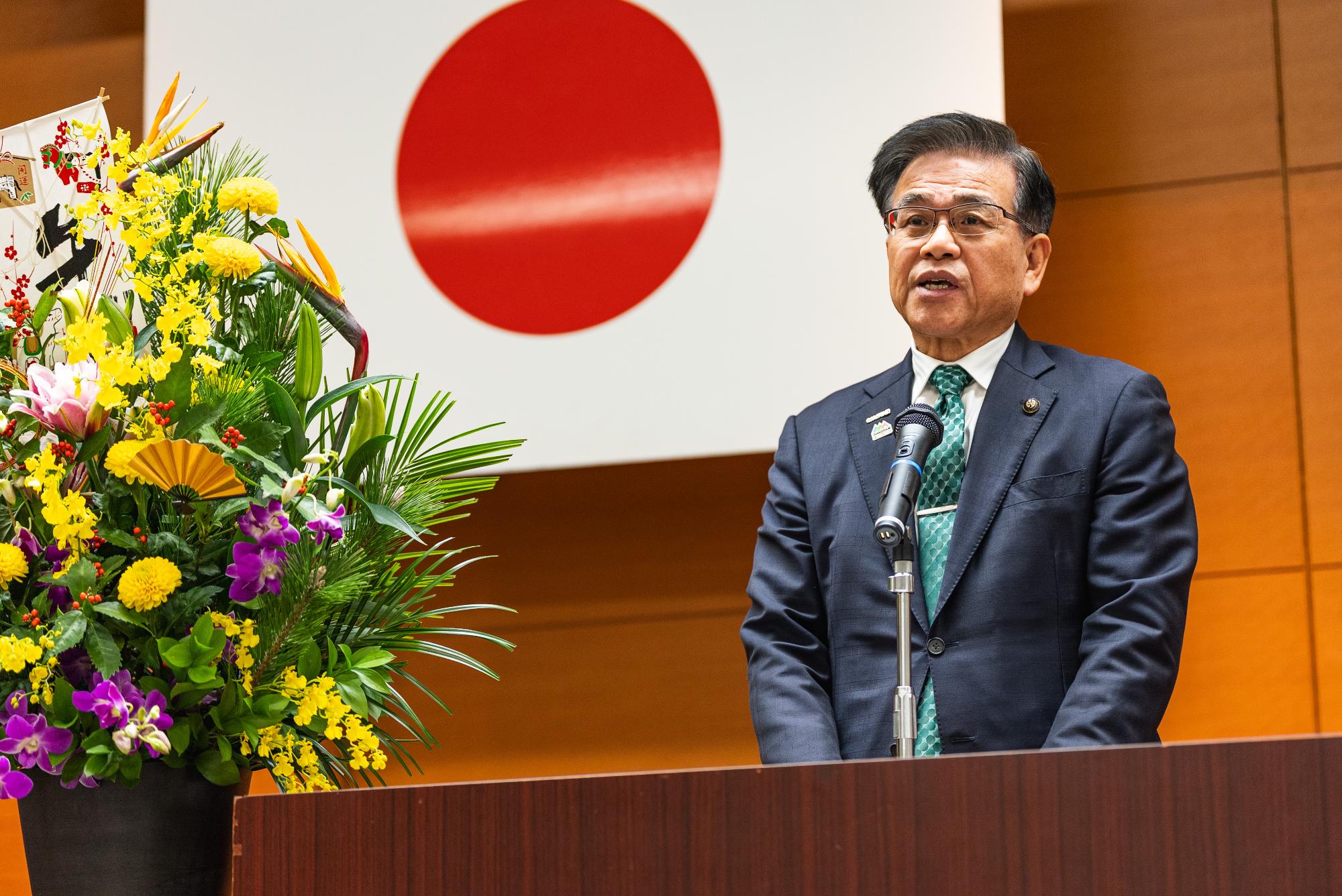 日の丸の旗の前で挨拶する市長。左側に花が飾られている。