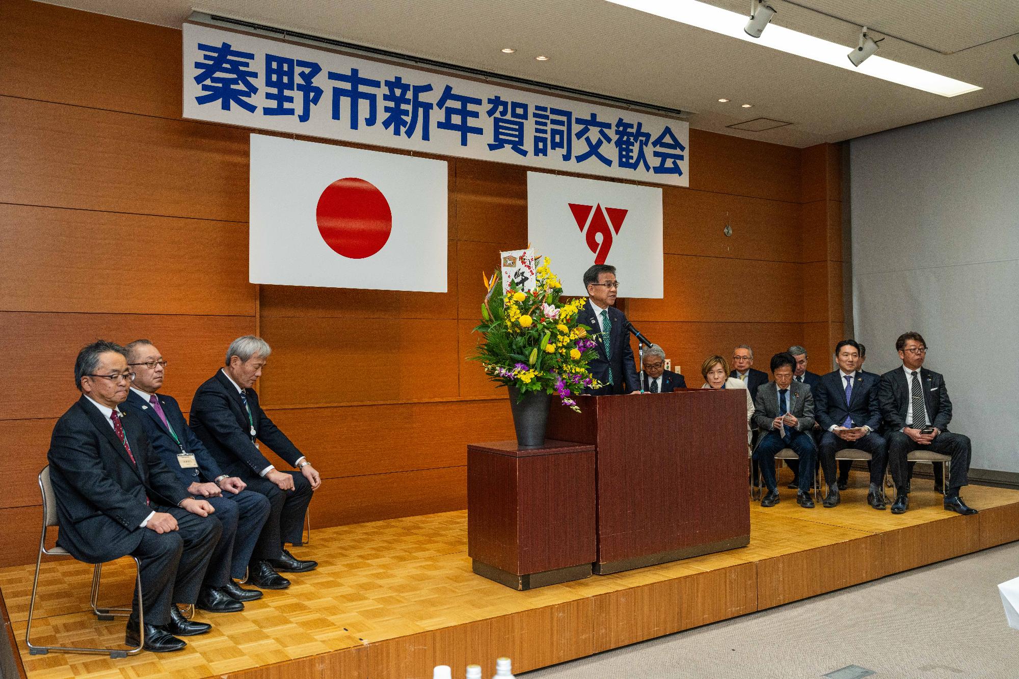 演台の後ろで挨拶する市長、右側に来賓10名が座り、左側に主催者3名が座る。