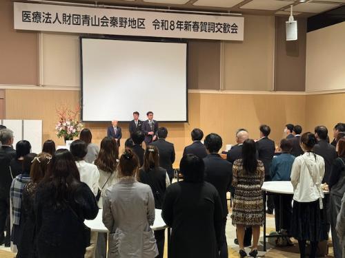 医療法人財団青山会秦野地区令和8年新春賀詞交歓会の看板とスクリーンの下で立つ市長と男性2人。手前には出席者の男女多数。