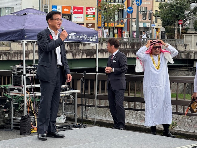 川沿いの道路上に張ったテントの前で挨拶する市長とその隣にアラブ系の衣装で仮装した男性