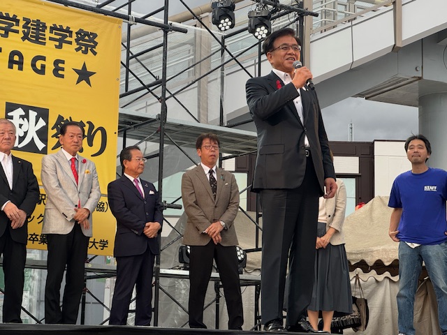 屋外のステージで挨拶する市長とその後ろに立つ来賓の男性4名