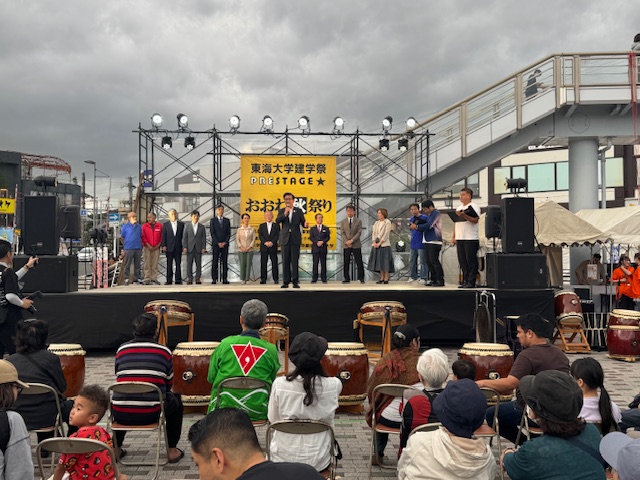 屋外ステージで挨拶する市長とその後ろに立つ多数の来賓、ステージ下には座って聞く市民、太鼓が置かれている