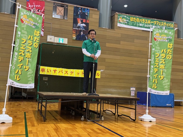 簡易ステージの上で挨拶をするTシャツ姿の市長、その左右はのぼり旗が置かれている