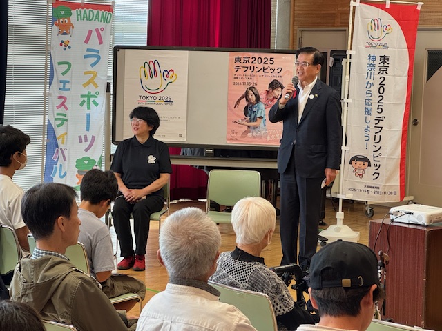 デフリンピックののぼり旗の横で挨拶する市長とそれを聞く椅子に座った多数の市民