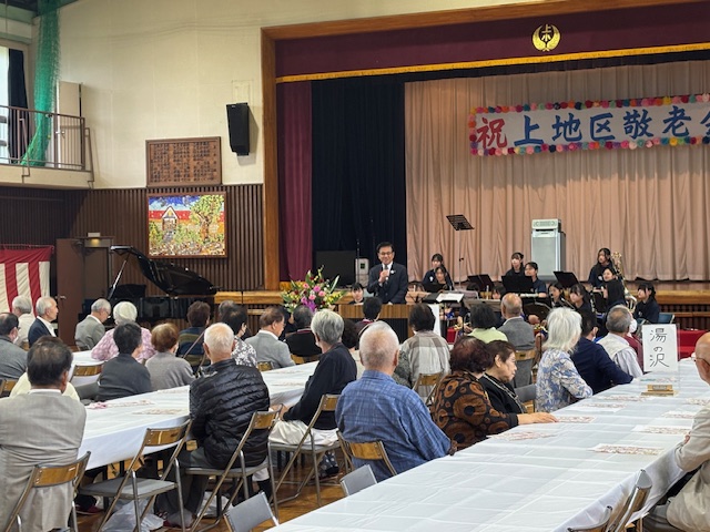 テーブルクロスのかかった長い机を囲んで座る高齢者を前に挨拶する市長