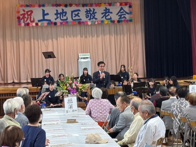 テーブルクロスをかけた長い机を囲んで座る高齢者を前に挨拶する市長