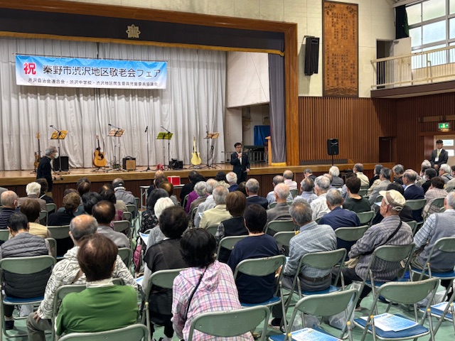 大勢の高齢者を前に挨拶する市長、ステージ上にはギターが3つ置いてある