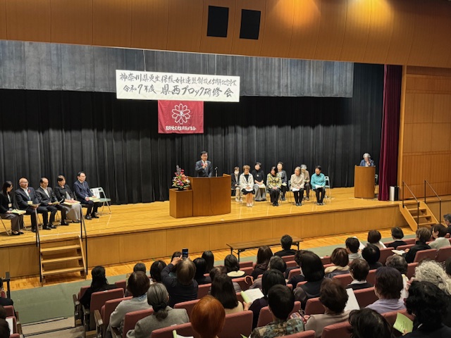 演台の後ろで挨拶する市長、その右側に座った女性8人、左側に来賓5人、その話を聞く多数の女性