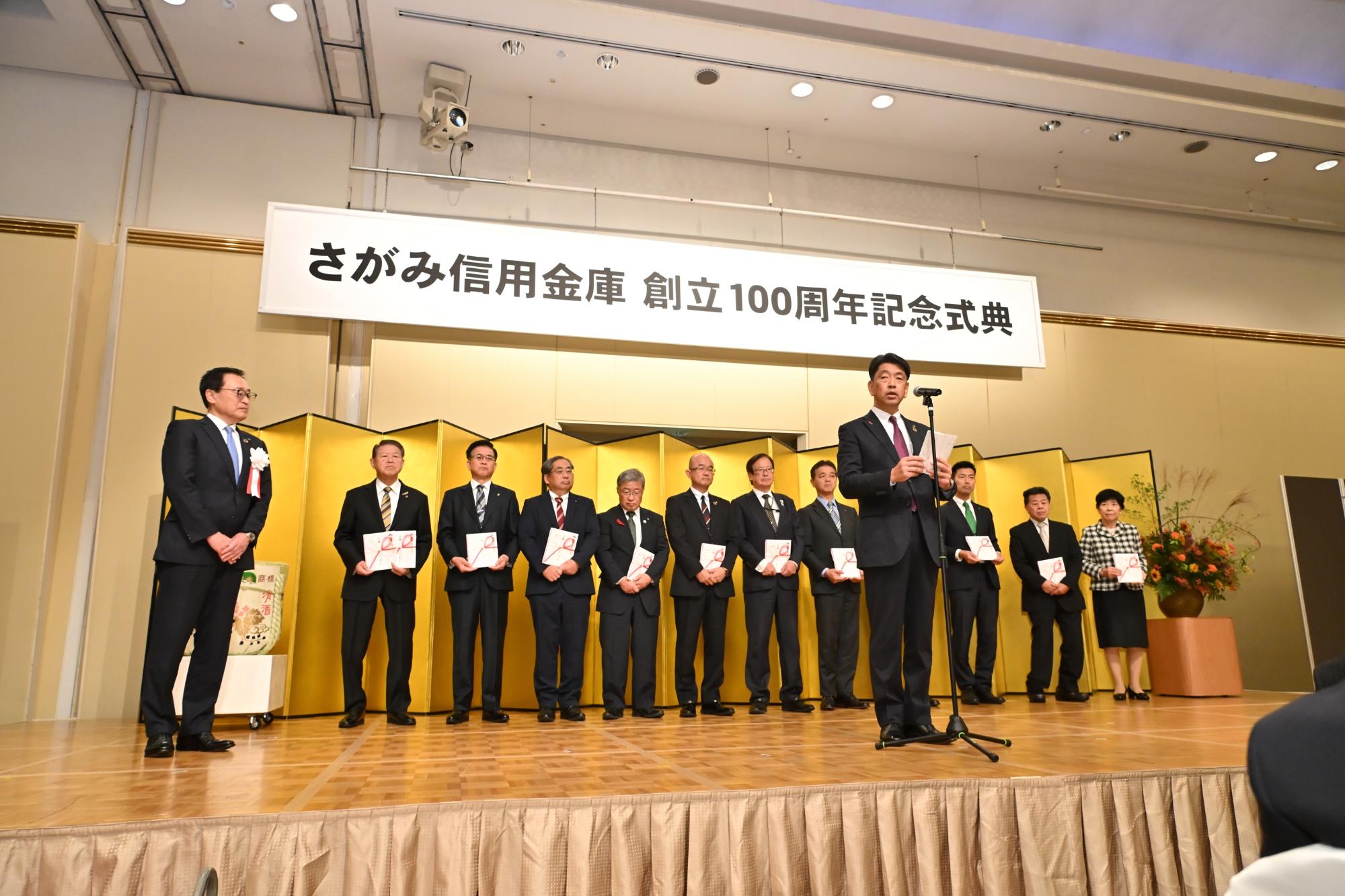 金屏風の前で整列する県西の市町長と挨拶をするスーツの男性