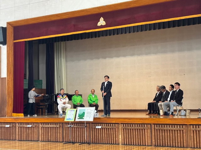 壇上で立って挨拶する市長と7人の男性の来賓がハの字に座っている