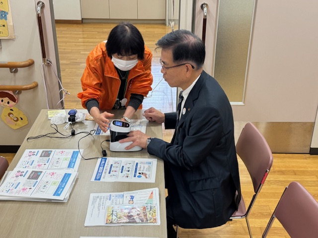 指を機械に入れて血管年齢を測定する市長と機会を操作する女性