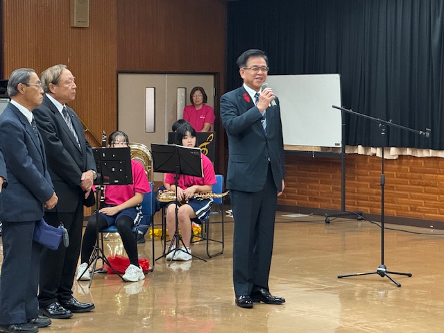立ってあいさつする市長とそれを聞く2人の男性の後ろに楽器をもった中学生たちが座っている