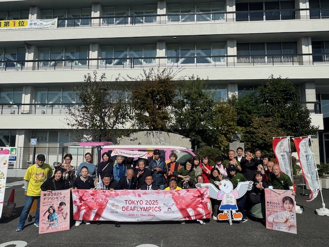ピンクのデフリンピックキャラバンカーの前で市長とデフリンピックを応援する大勢の市民の記念写真