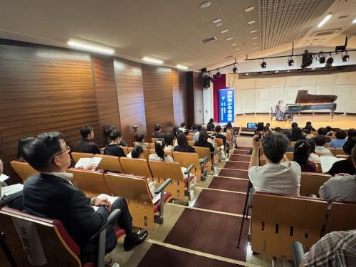 遠くのステージでピアノを弾く人の演奏を見学する市長