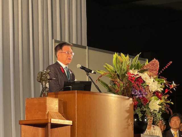 演台で挨拶する市長、左側には二宮尊徳の小さな銅像が置かれている。右側には大きな花が飾られている。