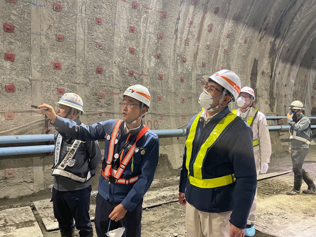 トンネルの中で作業着姿の男性が3人、1人が指をさして説明している。