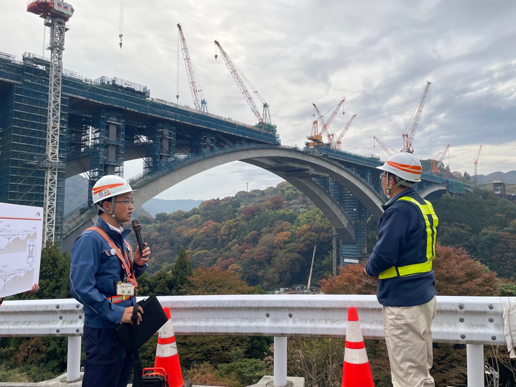 大きな橋を建設中の工事現場の前で作業着姿の男性が2人向かい合って話をしている