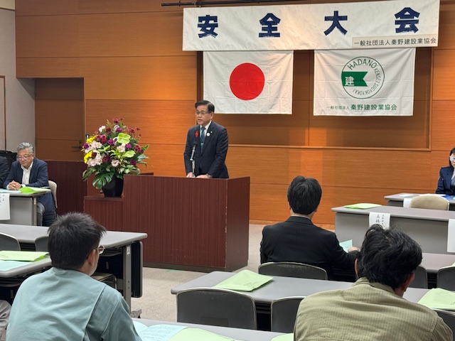 演台の後ろで挨拶する市長、頭上に安全大会の看板がある。手前に出席者の作業着姿の男性数人