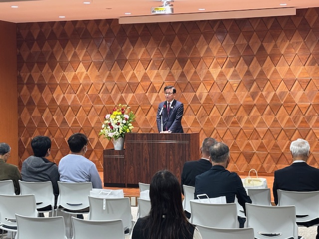 演台の後ろで挨拶する市長、その前方に受賞者が白いいすに7人座っている