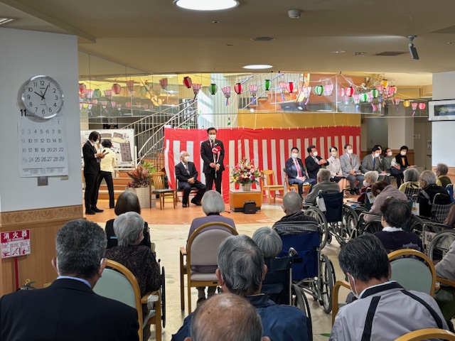 紅白幕の前で挨拶する市長とそれを聞く多数の椅子や車いすに座った高齢者
