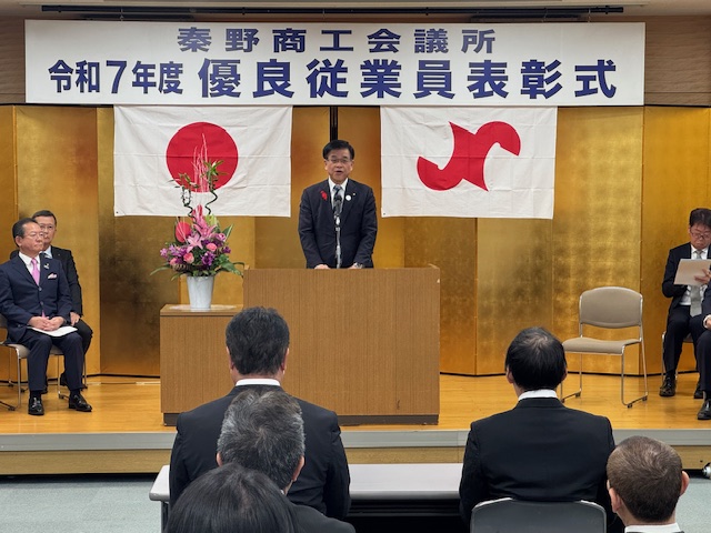 演台の後ろで挨拶する市長、頭上に優良従業員表彰式の看板とその下に国旗と市旗が掲げられている。