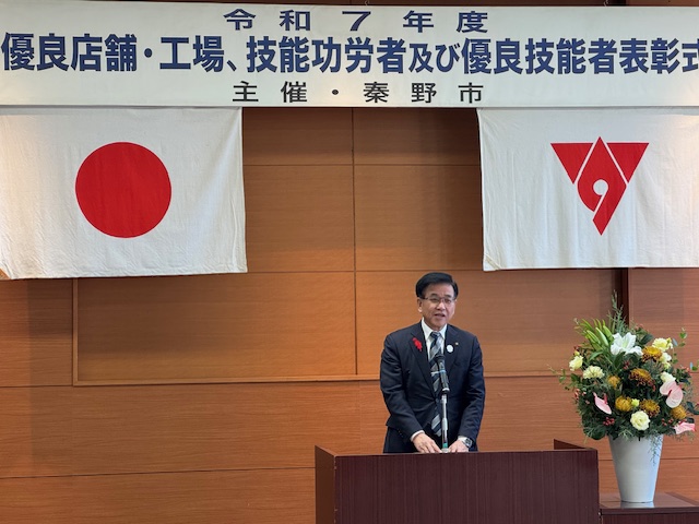 演台の後ろで挨拶する市長、頭上に優良店舗等表彰式の看板とその下に国旗と市旗が掲げられている。