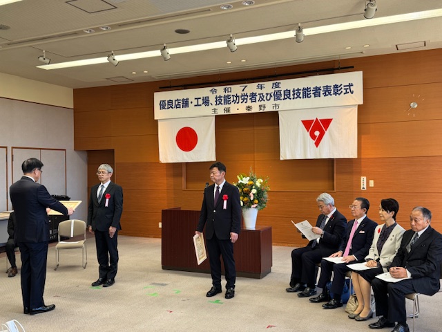 受賞者を前に賞状を読み上げる市長と、すでに受け取った男性、その横に来賓4名が座っている。