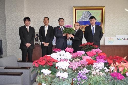 市長と一緒にシクラメンの鉢植えを持つ男性とその左右に人の男性、手前のテーブルの上は託宣のシクラメンで覆われている。