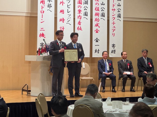 演台の前で感謝状を持つ市長と男性、その右側に来賓の男性3名が座る