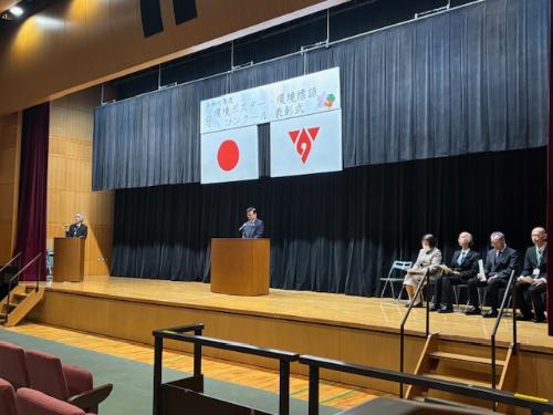 ステージの演台の後ろで挨拶する市長、奥には司会の男性、手前は椅子に座った来賓4人