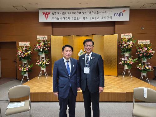 秦野市・坡州市海外友好都市20周年記念パーティーの看板のある部屋で市長とスーツの男性が立っている。背後にスタンド花が4基ある