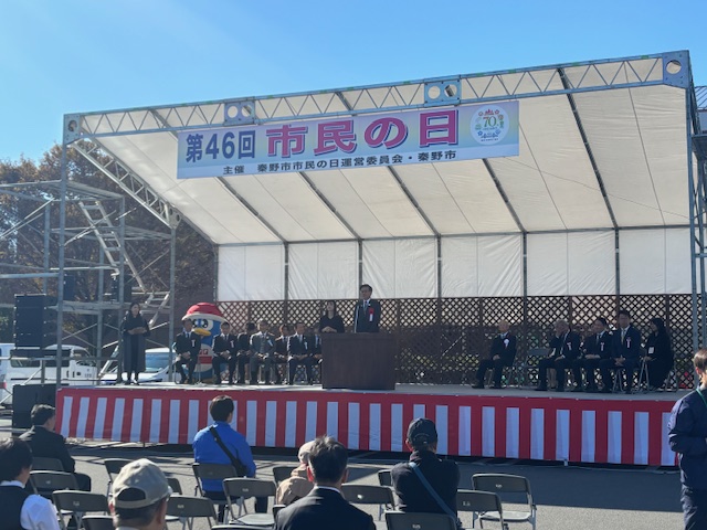 紅白幕で縁取られたステージの上に立ち挨拶する市長とその左右に多数の来賓が座る。手前に来場者10名程度が椅子に座っている