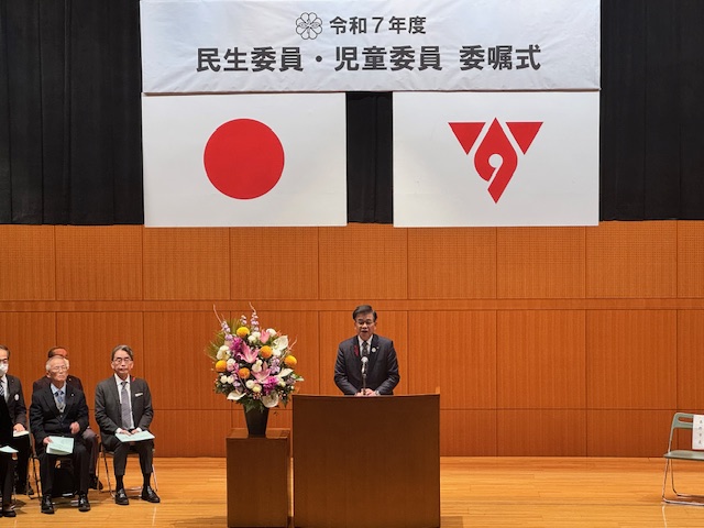 演題の後ろに立って挨拶をする市長、背後に国旗と市旗、左側に盛花が置かれている