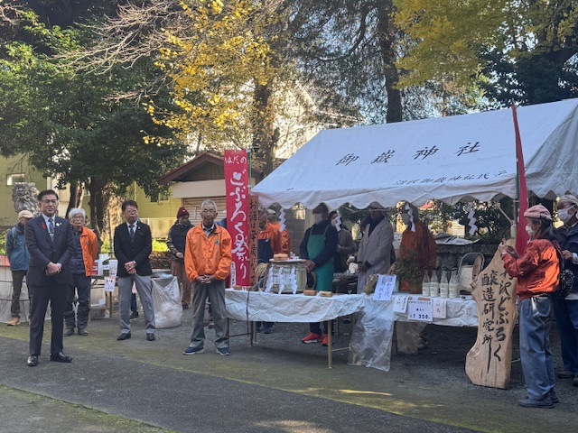 屋外のテントの前で挨拶をする市長と周辺に役員の男性多数