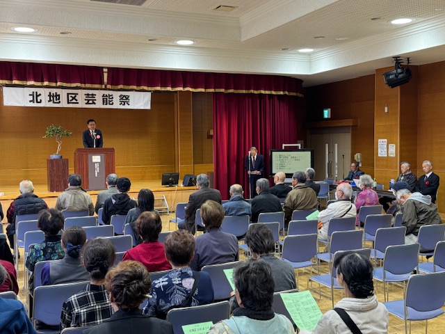 ステージの上で挨拶する市長とそれを聞く椅子に座った多数の高齢者、手前の数人は緑色のパンフレットを持っている