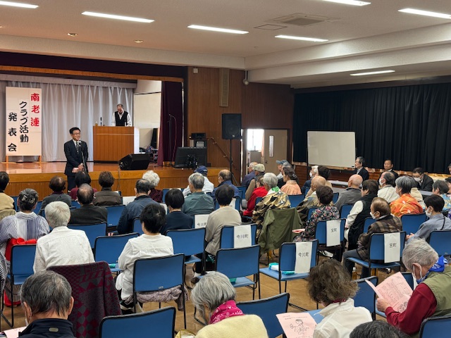 大勢の高齢者が座る奥で挨拶する市長。来場者はピンク色のパンフレットを持っている