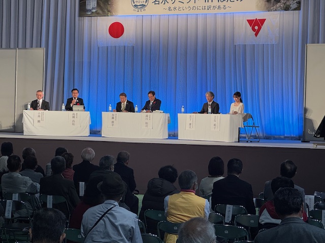 白いクロスのかかった机3台に2人ずつ座り、意見交換を行っている。ステージ下には椅子に座った観客多数。