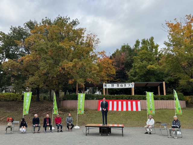 紅白幕と里山まつりののぼり旗、横断幕のある屋外で挨拶する市長と来賓が左に6人、右に2人座る