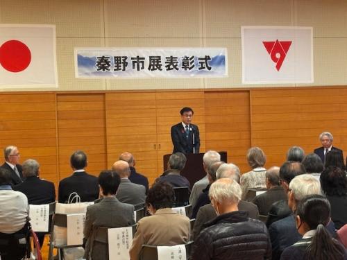 挨拶する市長の頭上に秦野市展表彰式の看板がある。手前に多数の受賞者が座る。