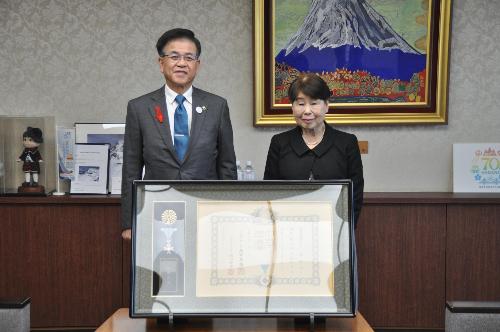 富士山の絵の前で、市長とスーツの女性が立っている。その前に賞状と勲章が入った大きな額縁が置いてある。
