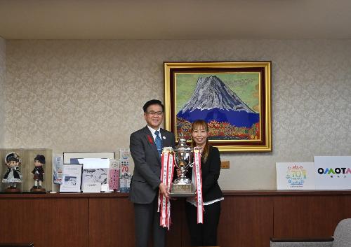 市長とスーツの女性が大きなトロフィーを持って立っている。