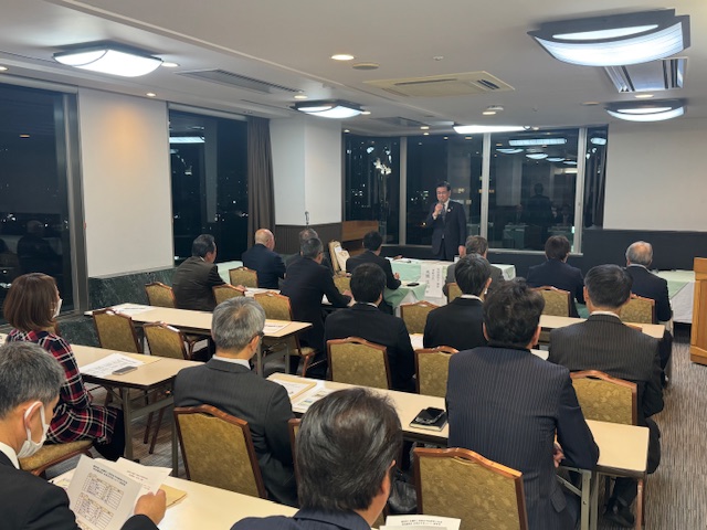 長机に2、3人ずつ座ったスーツの男性が20人ほどいる奥に市長が挨拶をしている。